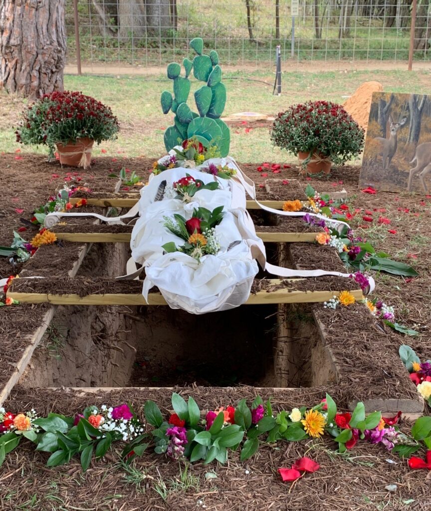 A shrouded burial laid over an open grave edged with flowers and potted plants
