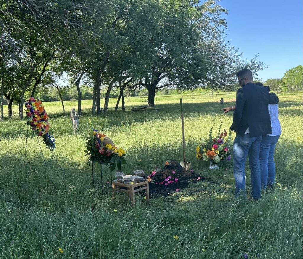 Two people standing near a grave decorated with flower arrangements in a green pasture