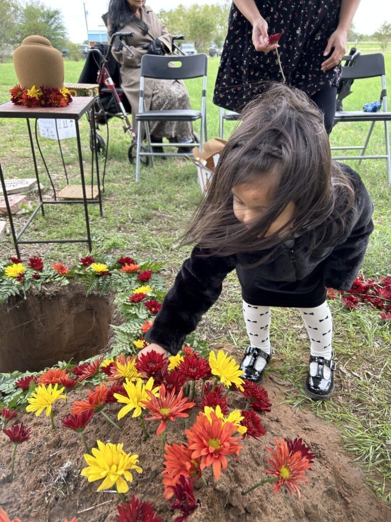A young girl placing a flower on a mound of soil covered in colorful flowers, with an urn on a table nearby